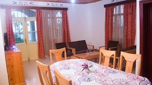 A dining area with a table set for four, wooden chairs, a small table with a laptop, and a view through a window with curtains. - Rebero Kivu Hotel (Bwishyura)