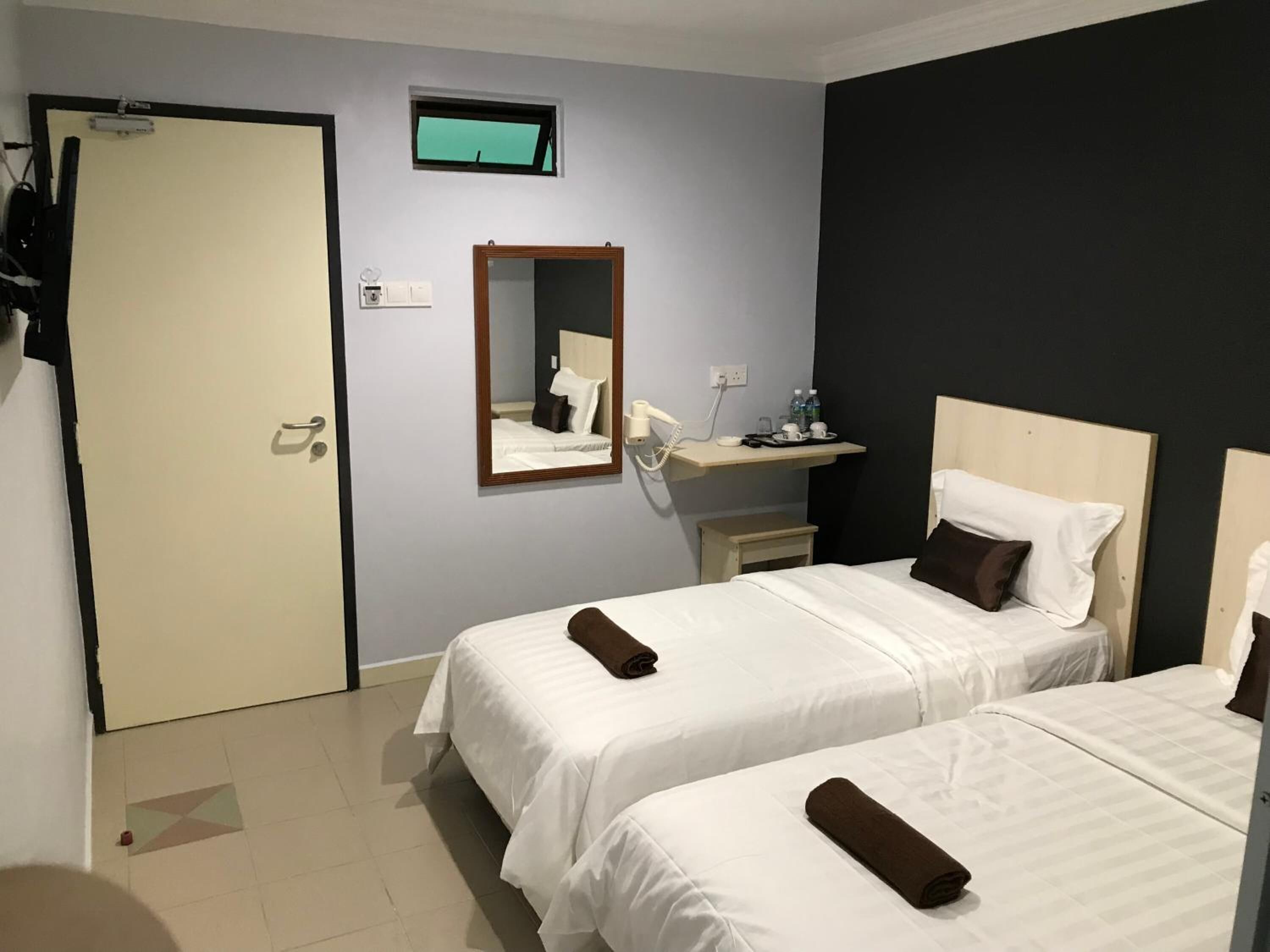 A hotel room with two beds, a small bedside table, a mirror, a shelf with toiletries, and a door.
