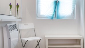 Een kamer met een witte stoel, een witte plank met twee paarse tulpen en een raam met blauwe jaloezieën.