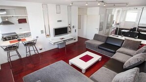 A modern living room with a grey sofa, a white coffee table, and a dining area with a white table and two bar stools. - Apartment Sunset (Belgrade)
