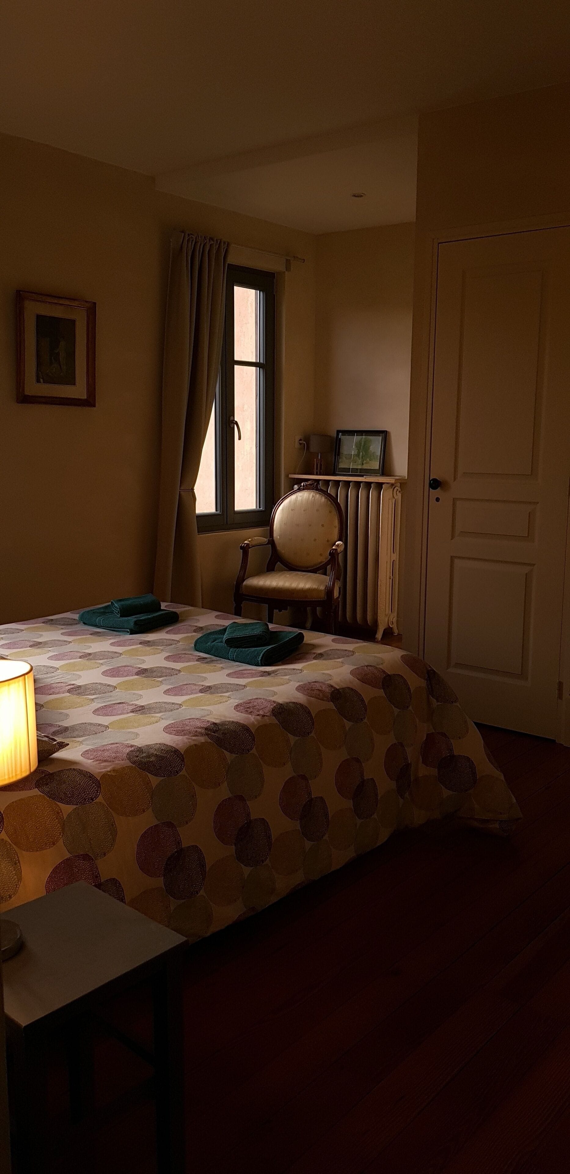 A room with a bed, a chair, a table, a lamp, a window with curtains, and a door.