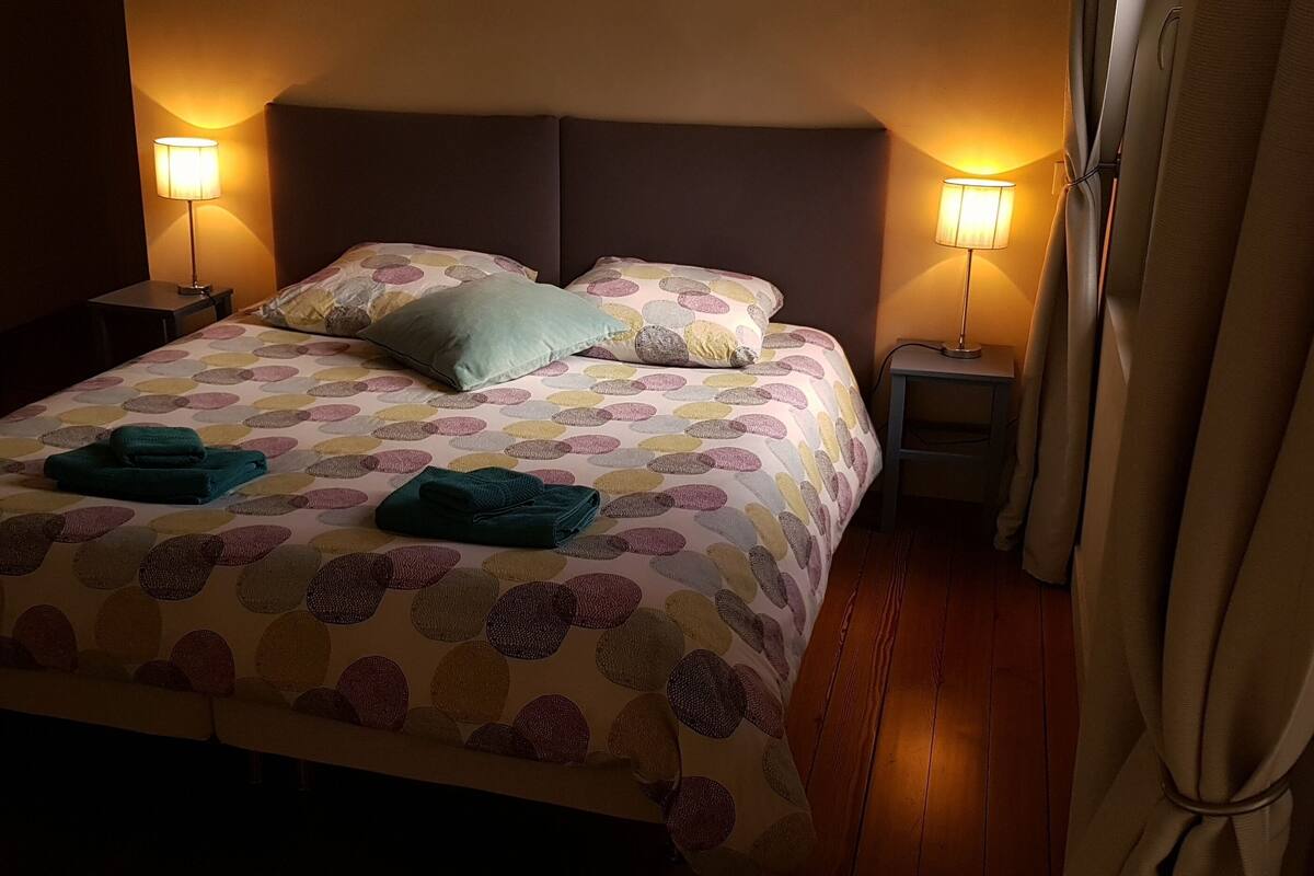 Een net opgemaakt bed met een geborreld sprei, twee kussens en twee opgevouwen handdoeken. Twee nachtlampjes en een nachtkastje met een lamp staan ernaast.