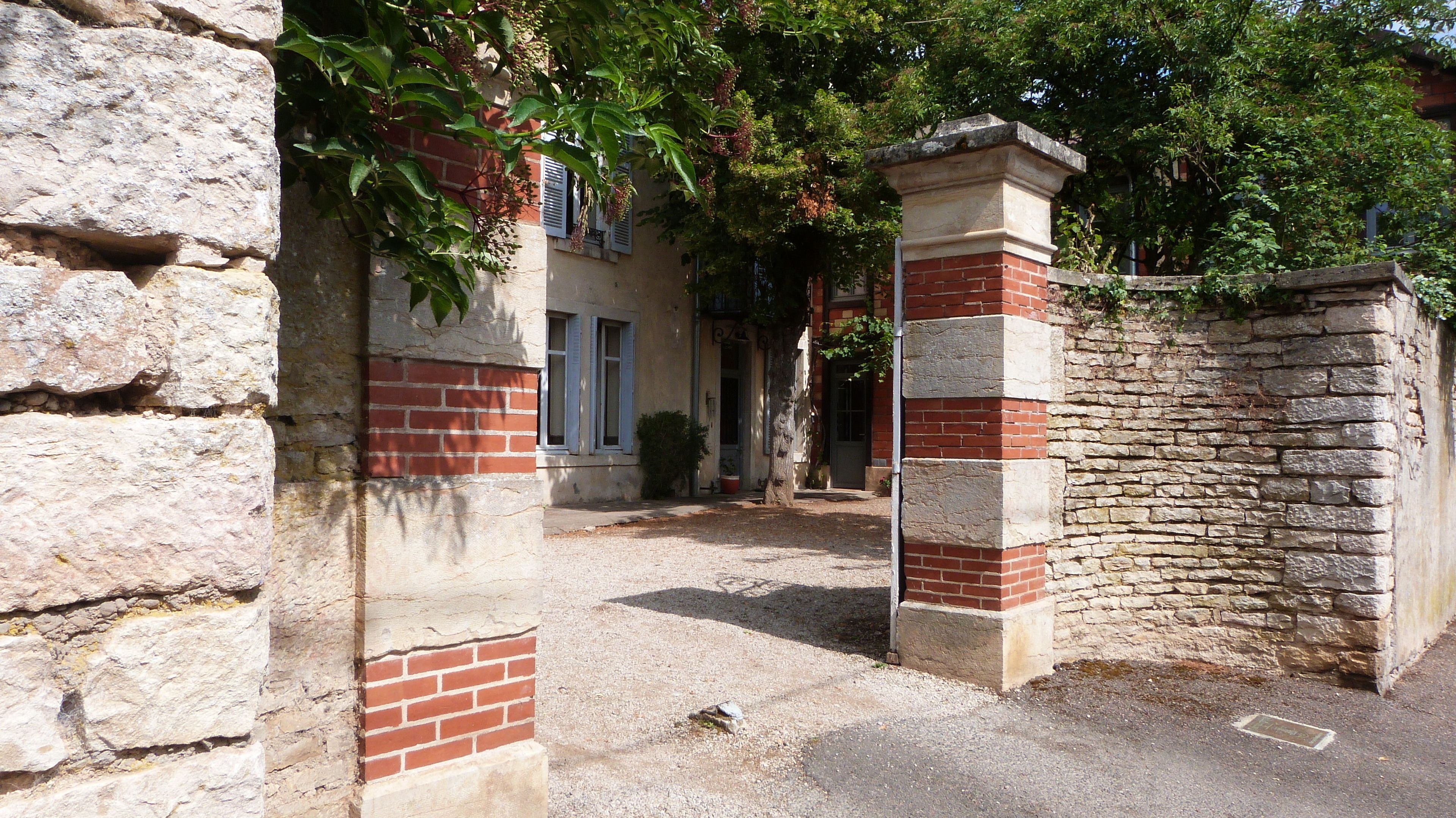 Un bâtiment en pierre avec une façade en briques, une cour recouverte de gravier et un arbre aux feuilles vertes.