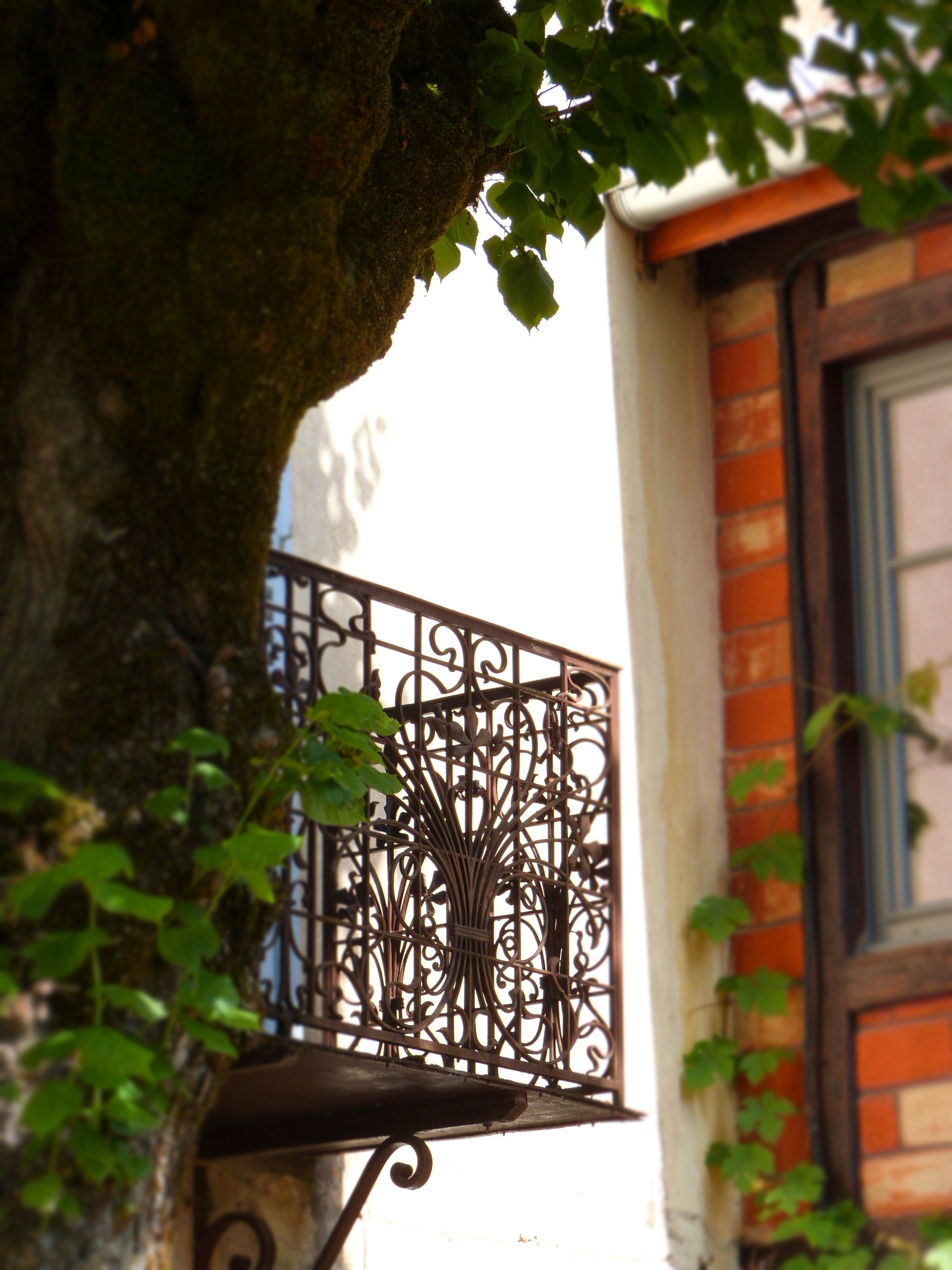 A wrought iron balcony with intricate designs, a brick wall, and a window with blue shutters.