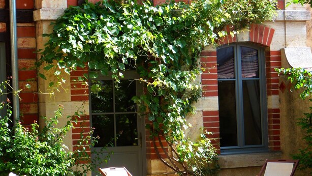 Un bâtiment à la façade en briques, doté de deux fenêtres et d’un mur recouvert de lierre. On y trouve deux chaises en bois et une table devant.