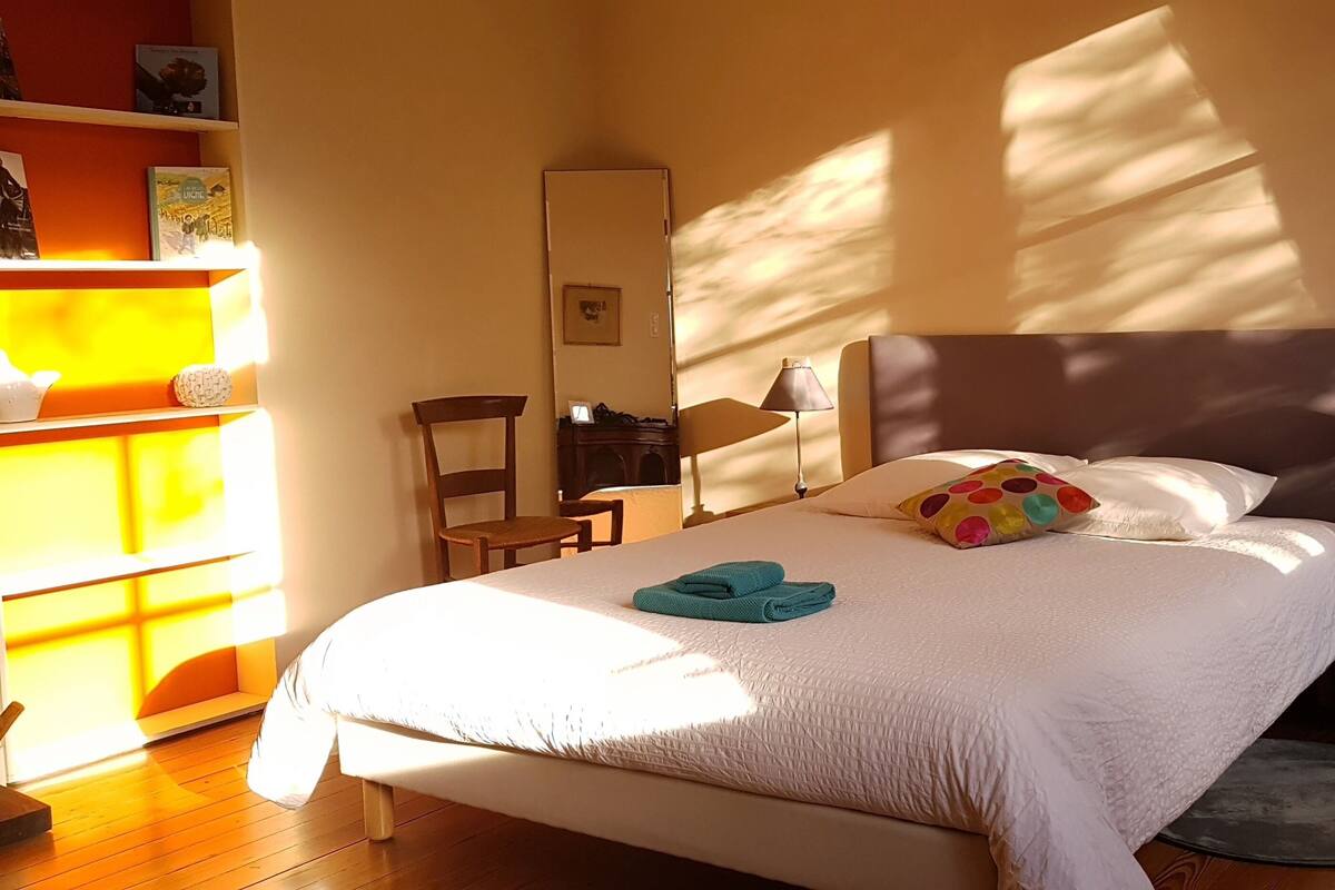 Een net opgemaakt bed met een witte deken en teal handdoeken, een bedlamp, een stoel en een boekenkast met boeken en decoratieve objecten.