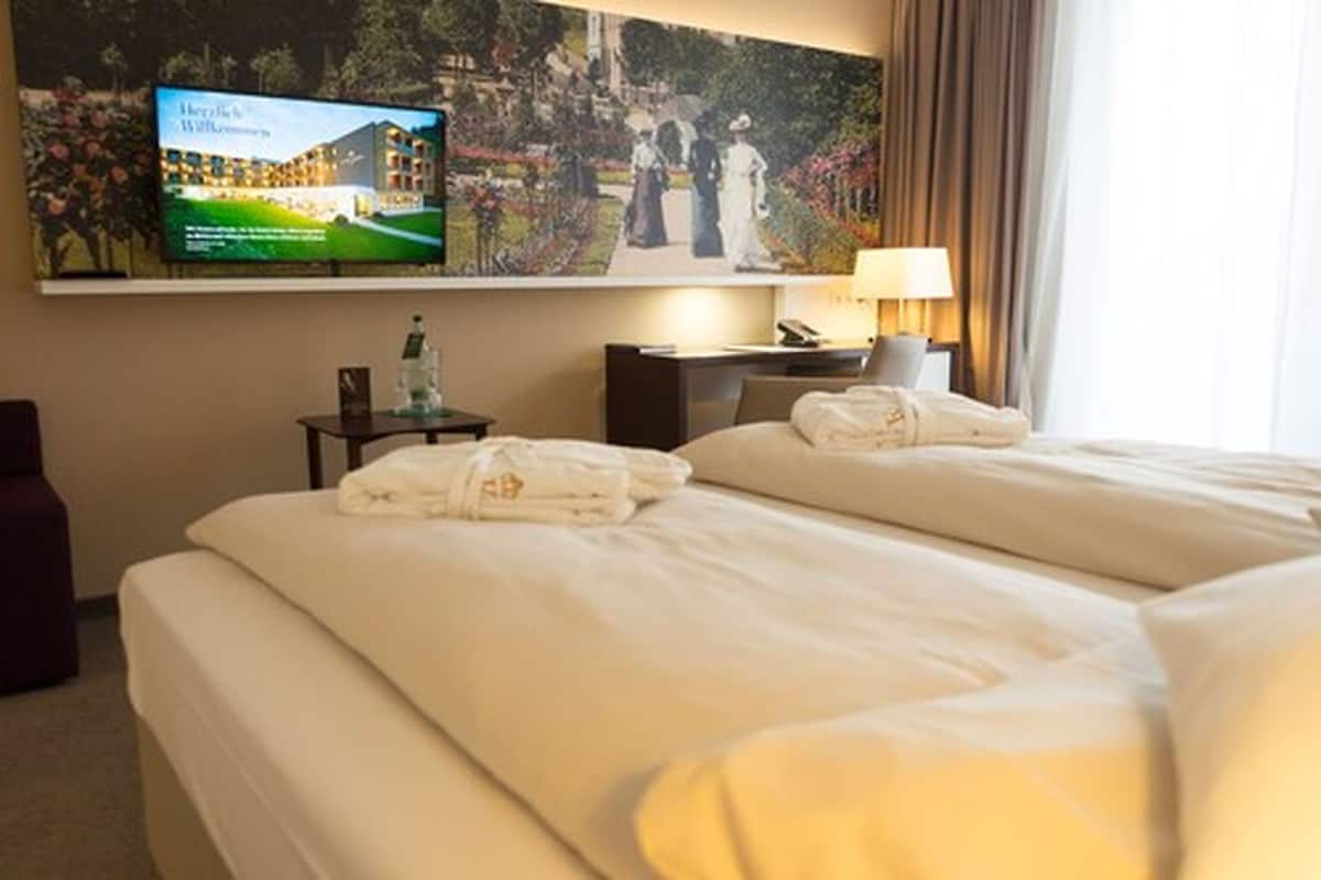 A hotel room with two beds, a television mounted on the wall, a desk, and a lamp.