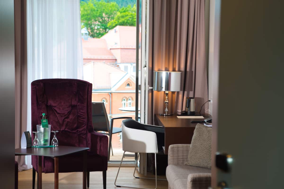A hotel room with a large window, a desk with a lamp, a chair, a sofa, and a small table with a bottle and glasses.