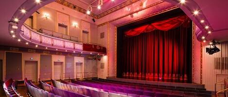 An auditorium with tiered seating, a stage with red curtains, and a balcony.
