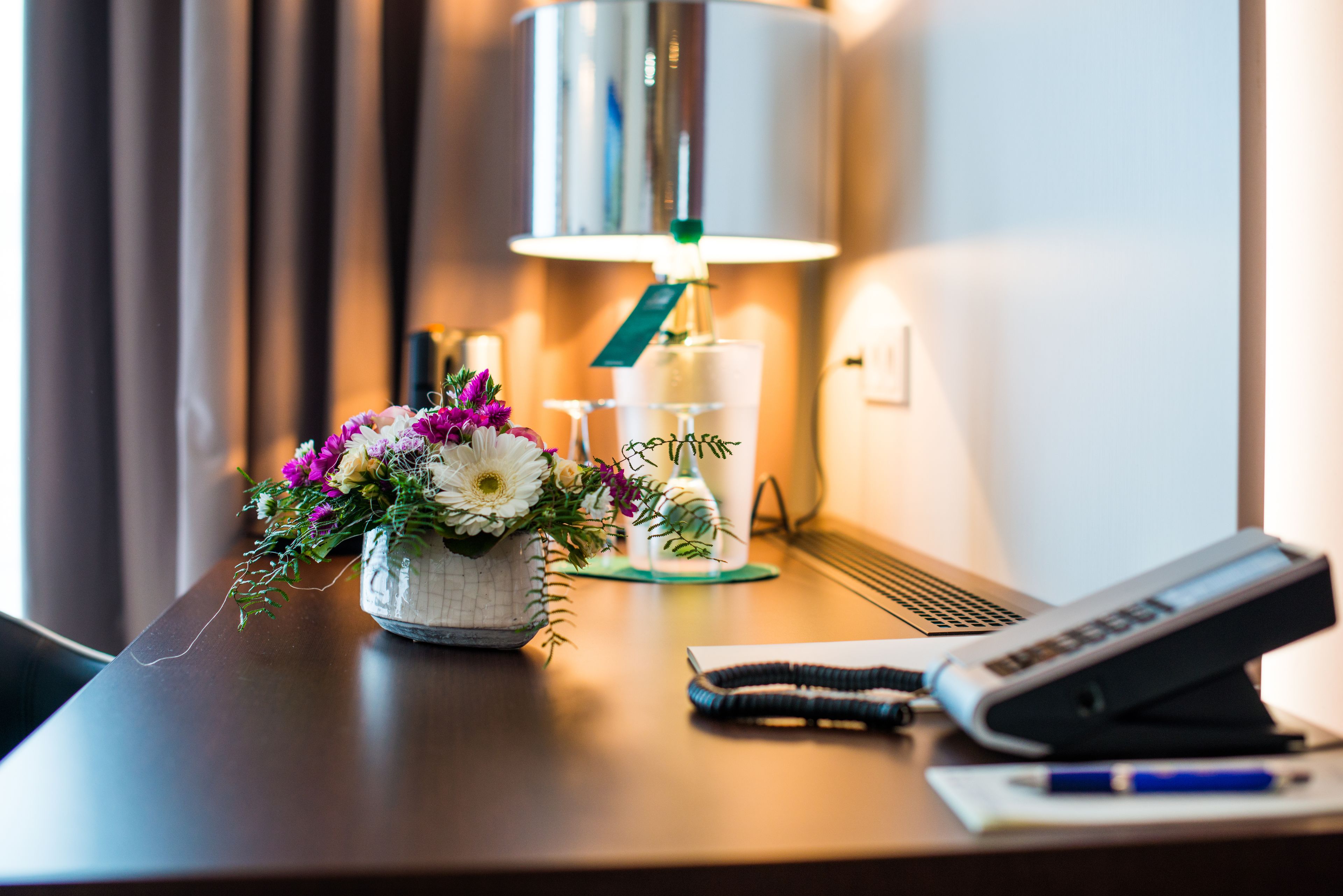 A desk with a lamp, a floral arrangement, a glass, a pen, and a phone.