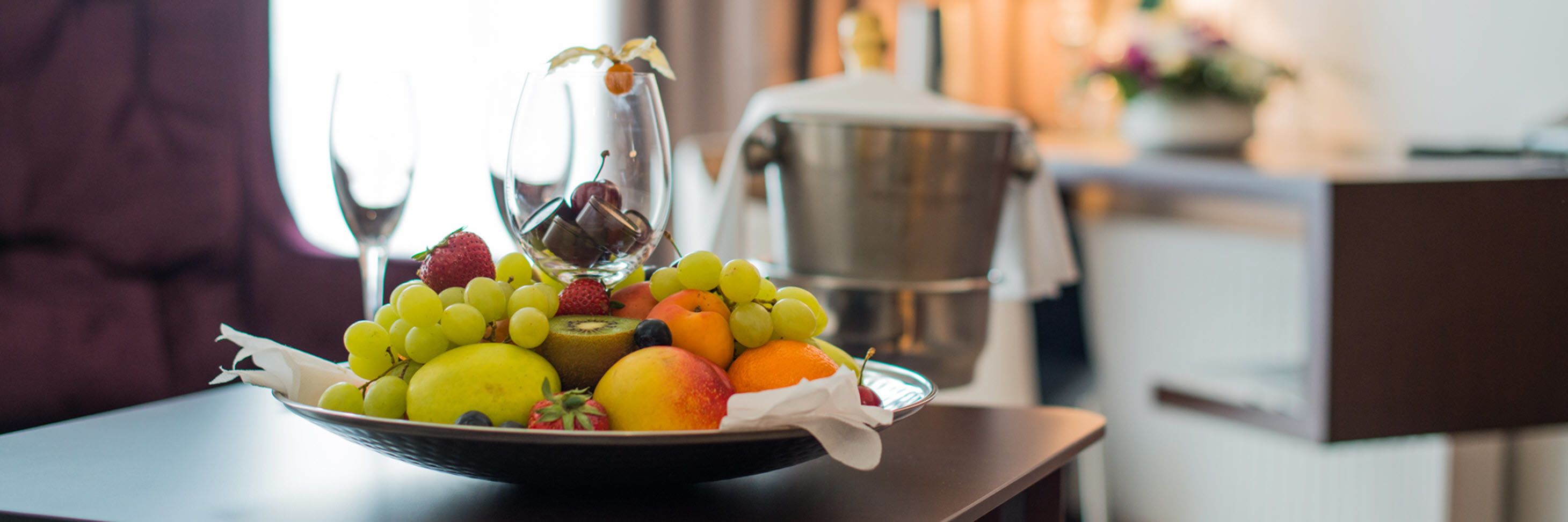 A fruit bowl on a table with a pitcher of water and a coffee maker in the background.