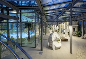 A modern indoor pool area with glass walls, lounge chairs, and a covered seating area. - Hotel König Albert (Bad Elster)