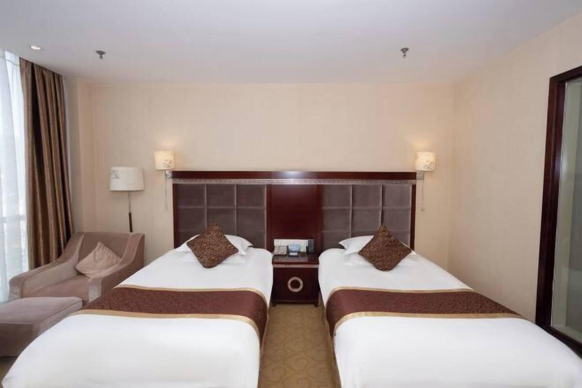 Ein Hotelzimmer mit zwei Betten, einem Nachttisch, einer Lampe, einem Sessel und einem großen Fenster.