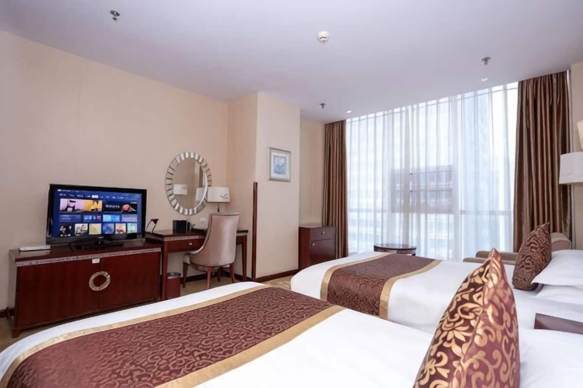 Ein Hotelzimmer mit zwei Betten, einem Schreibtisch mit Computer, einem Fernseher und einem großen Fenster mit Vorhängen.