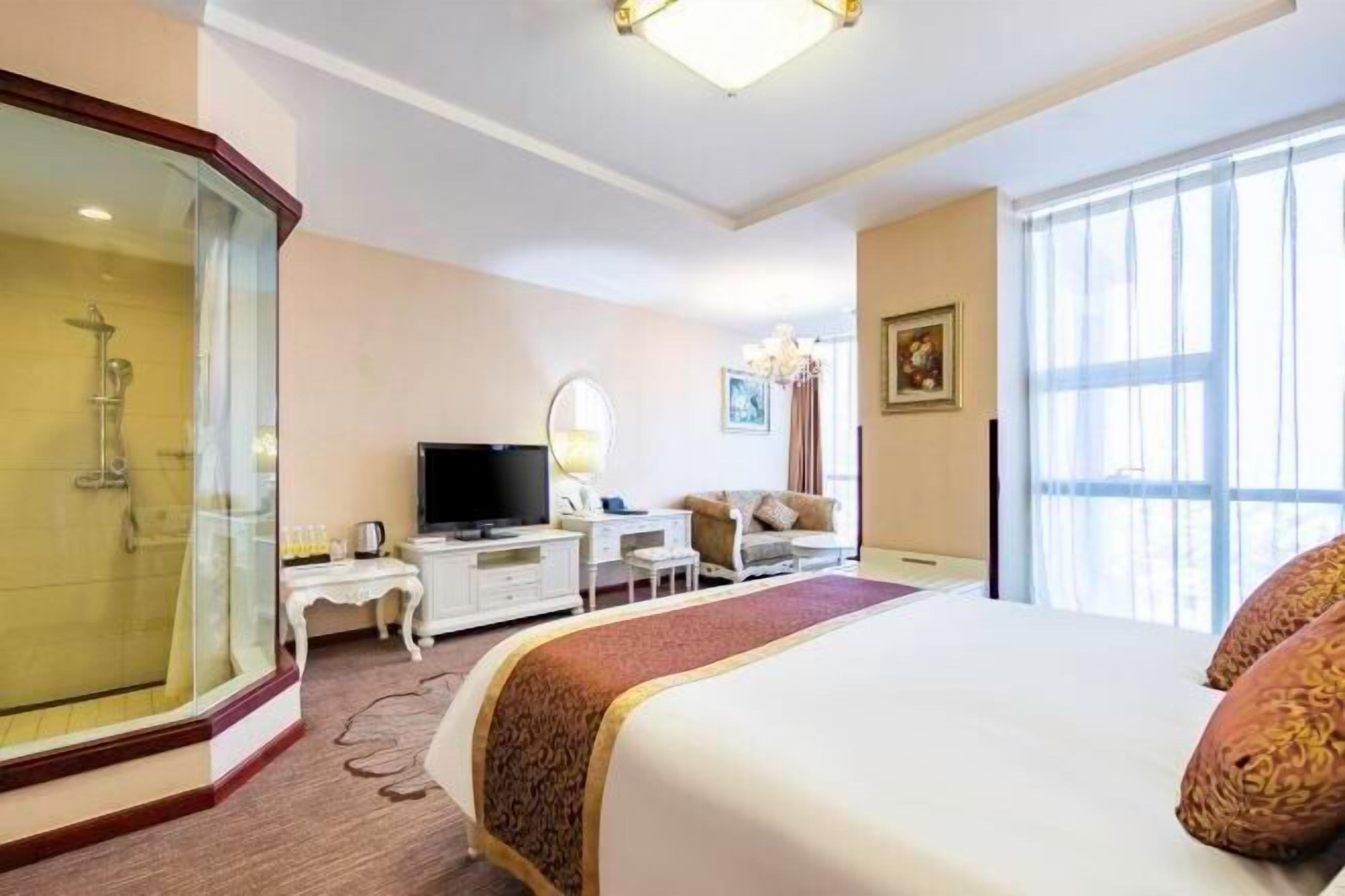 Quarto de hotel com cama, televisão, escrivaninha, cadeira, espelho e chuveiro.