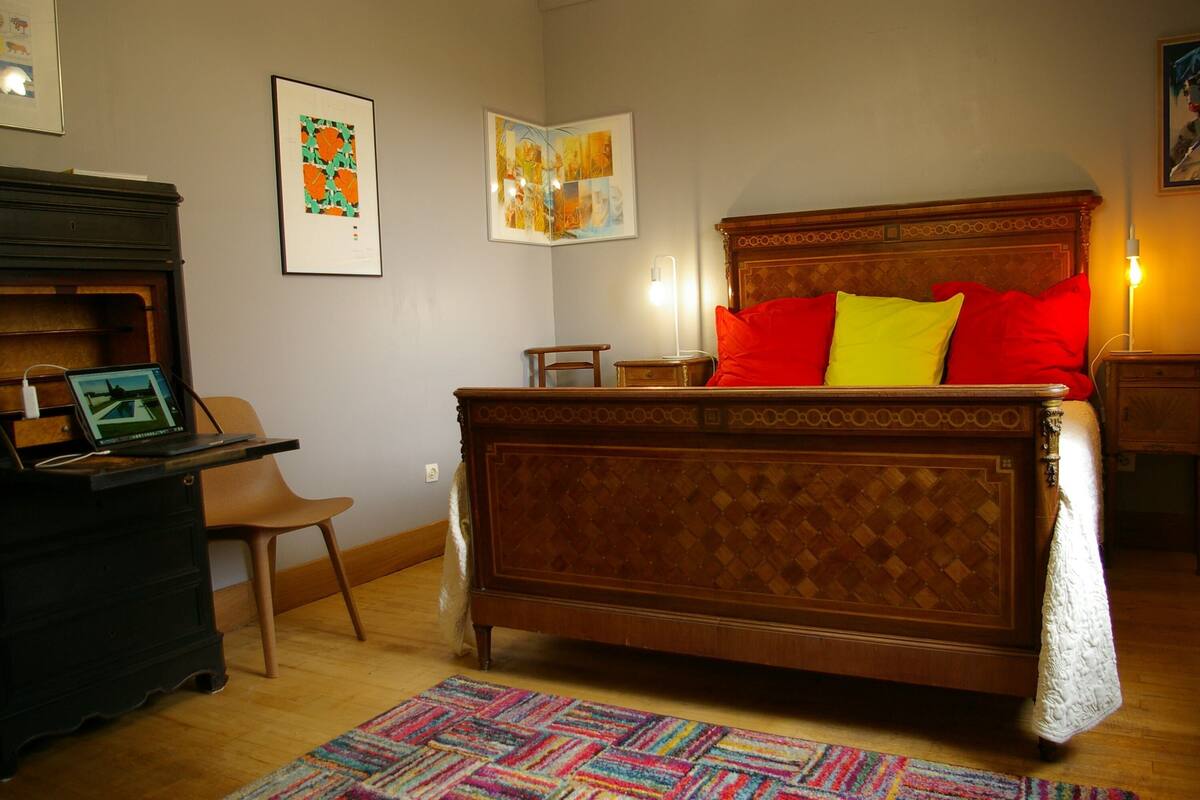 Une chambre avec un lit en bois, un bureau avec un ordinateur portable, une chaise et un tapis coloré.