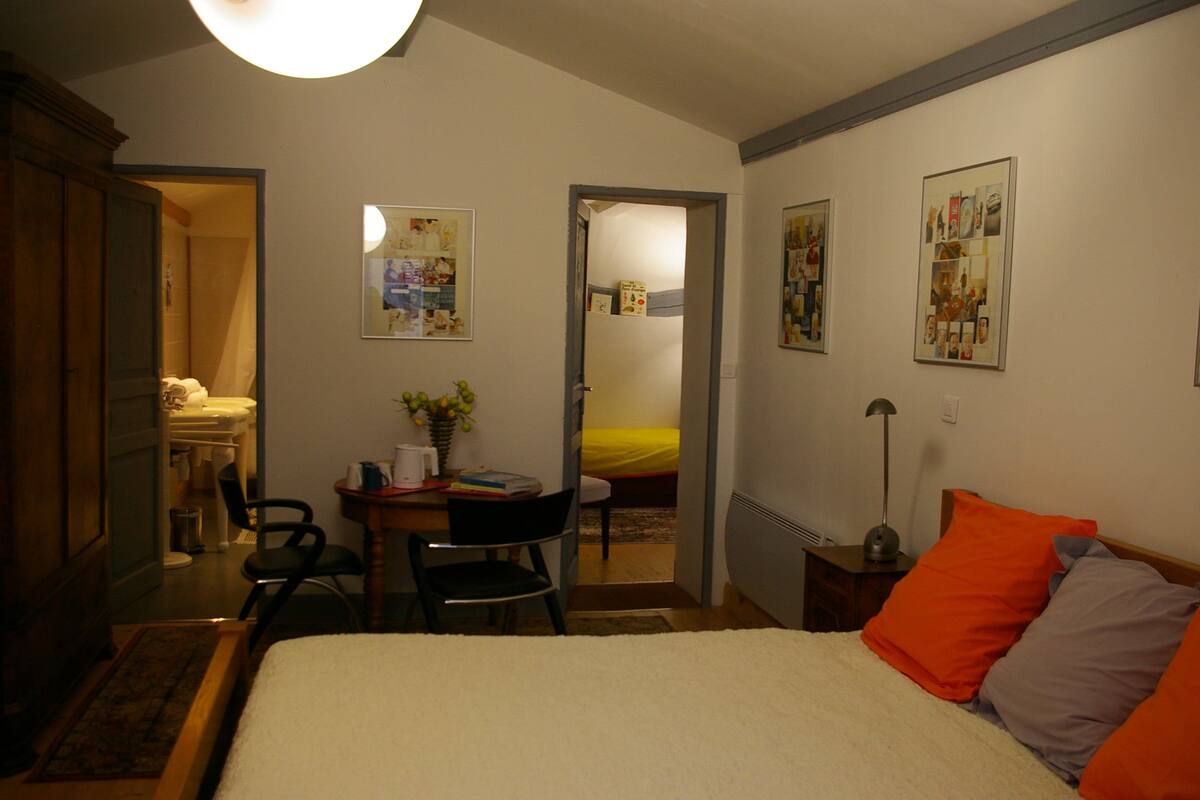 Une chambre à coucher comprenant un lit, un bureau, une chaise et une petite table sur laquelle est posé un vase de fleurs. Deux portes donnent accès à d’autres pièces.