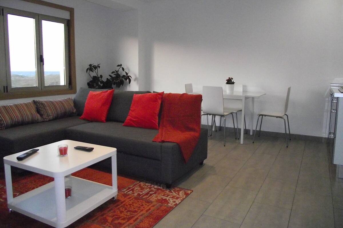 Una sala de estar moderna con un sofá gris, cojines rojos, una mesa de centro blanca y un comedor con mesa y sillas blancas.