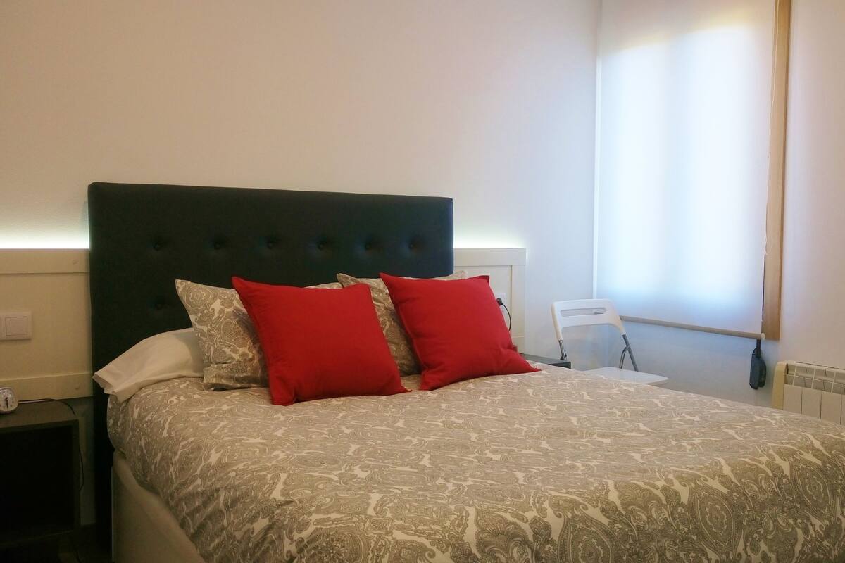 Una cama con una colcha estampada y dos almohadas rojas, un cabecero oscuro, una mesita de noche con un reloj y una ventana con persianas.