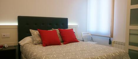Una cama bien hecha con una colcha estampada y dos almohadas rojas, un cabecero oscuro y una mesita de noche con un reloj y un vaso de agua.