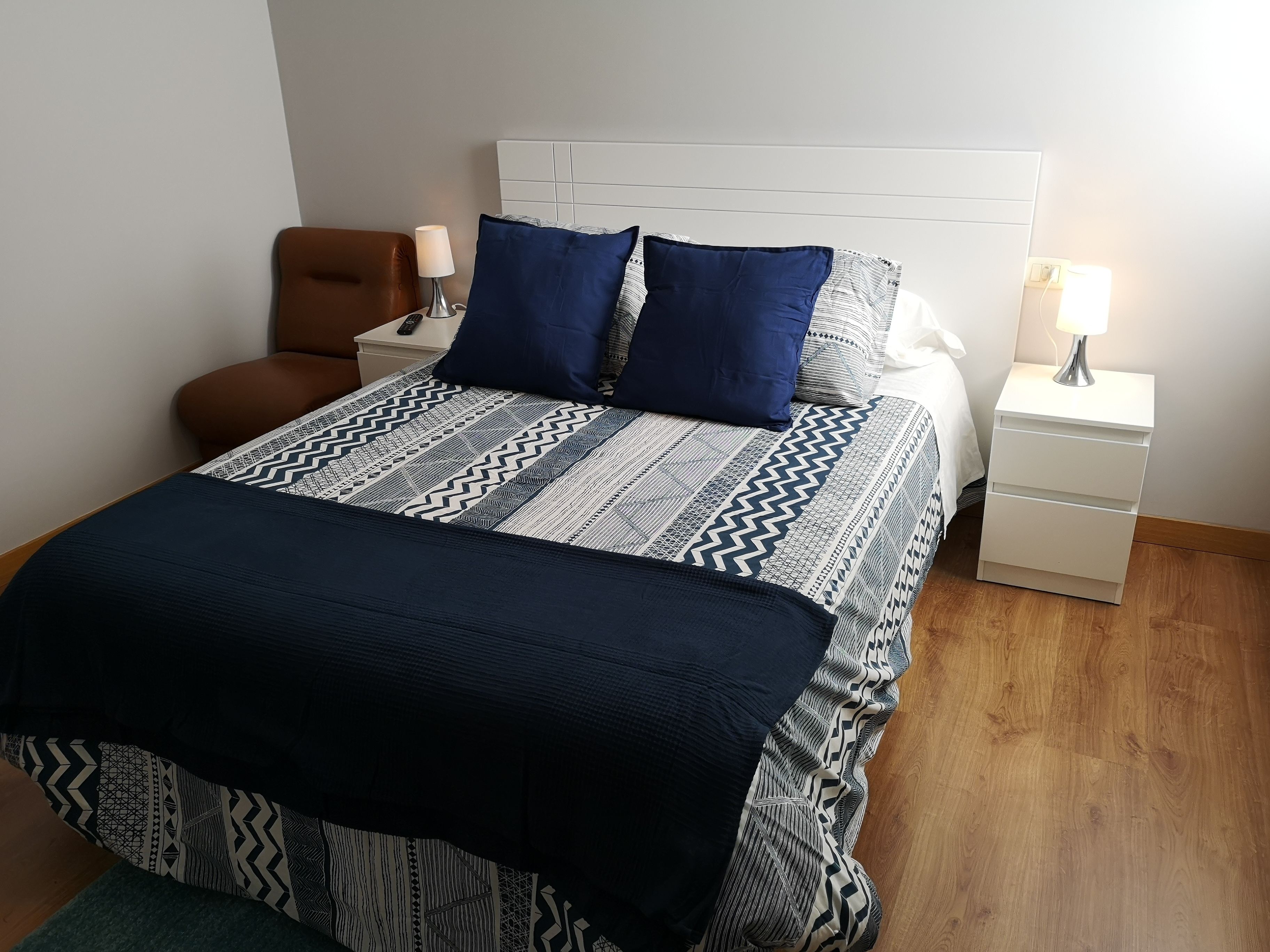 A neatly made bed with a patterned bedspread and pillows, a brown chair, and a white bedside table with a lamp.