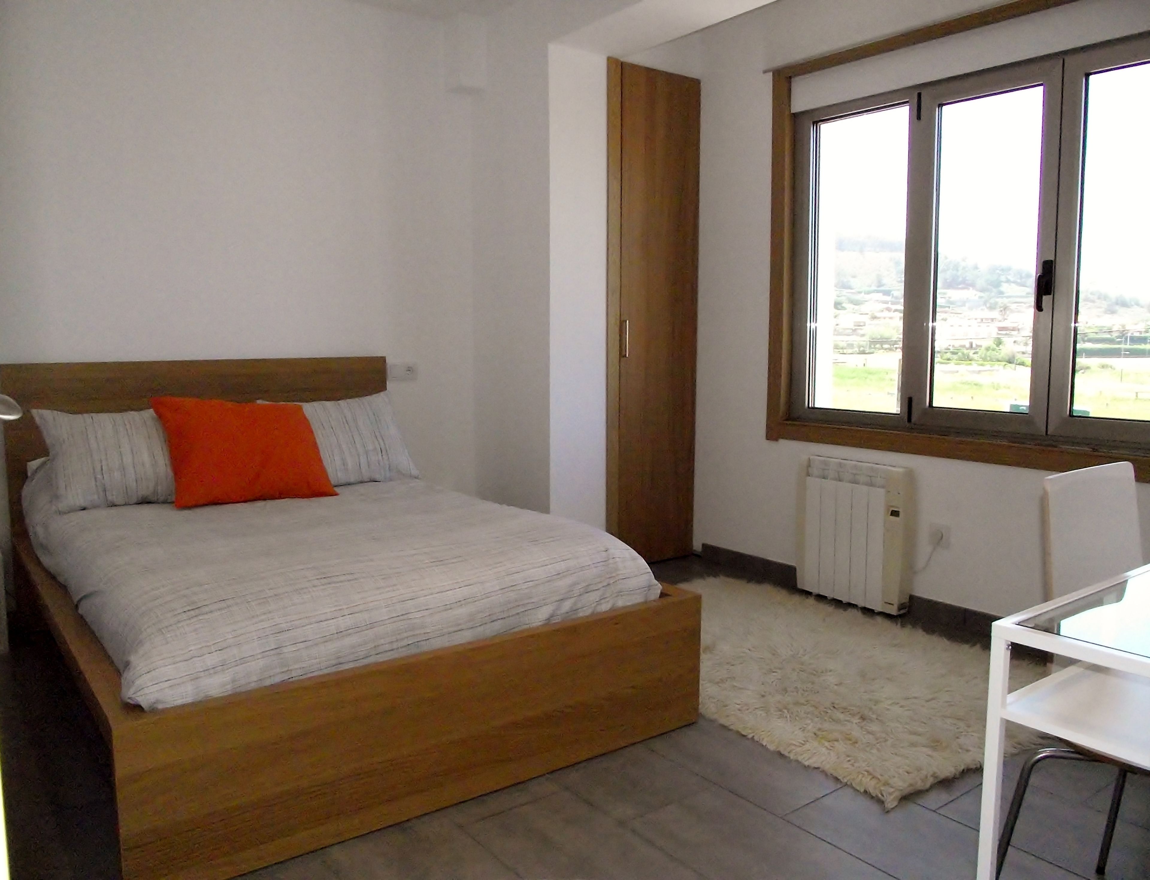 Une chambre à coucher avec un lit en bois, un radiateur blanc, une fenêtre donnant sur un beau paysage et un bureau blanc.