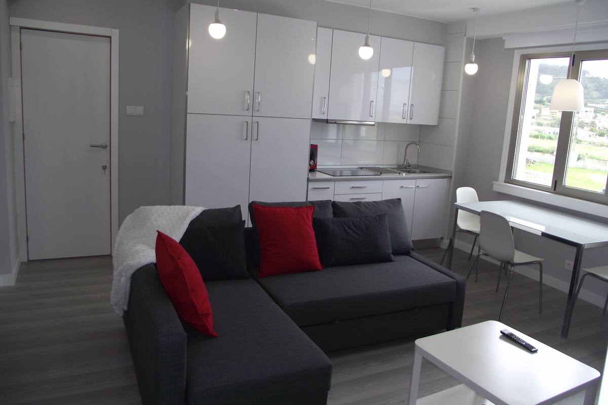 Una cocina moderna con armarios blancos, un comedor con una mesa y sillas blancas, y un sofá seccional gris con cojines rojos.