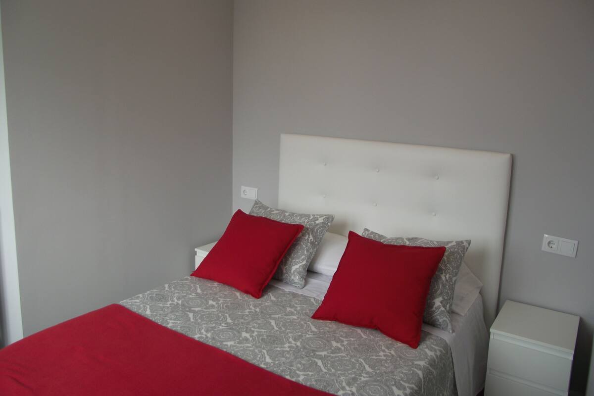 Un dormitorio con una cama, dos almohadas rojas, una colcha estampada, una mesita de noche blanca y una pared gris.