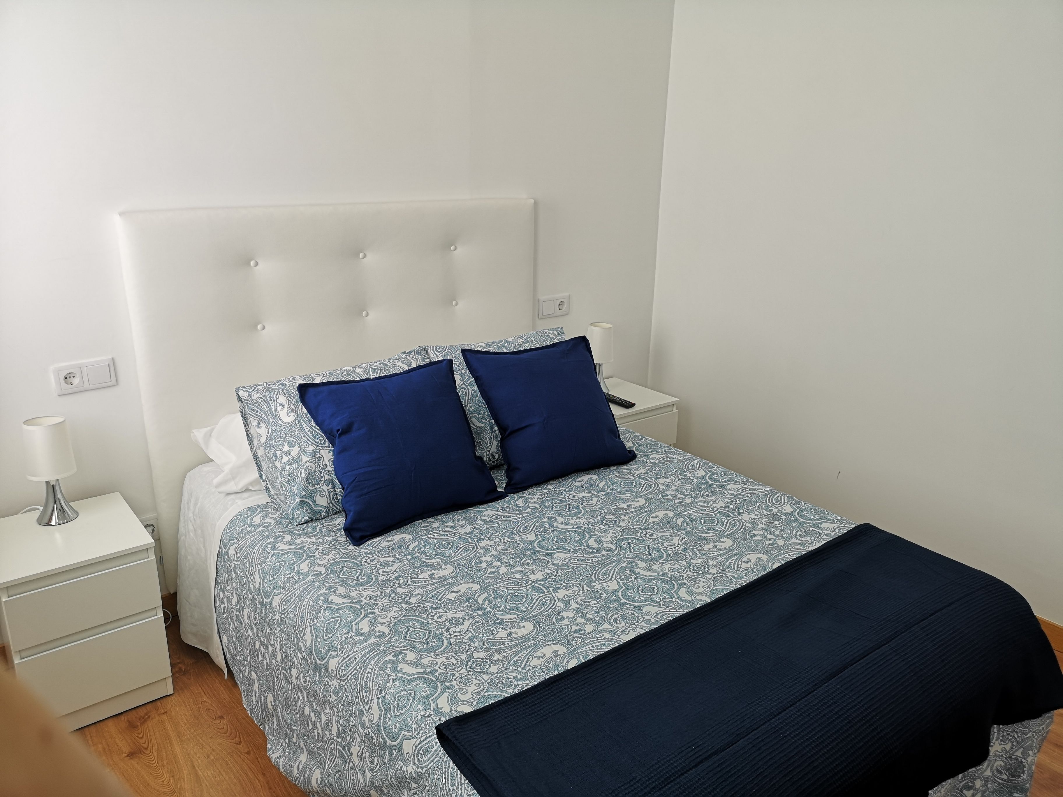 A neatly arranged bedroom with a patterned bedspread, two pillows, a bedside table with a lamp, and a nightstand.
