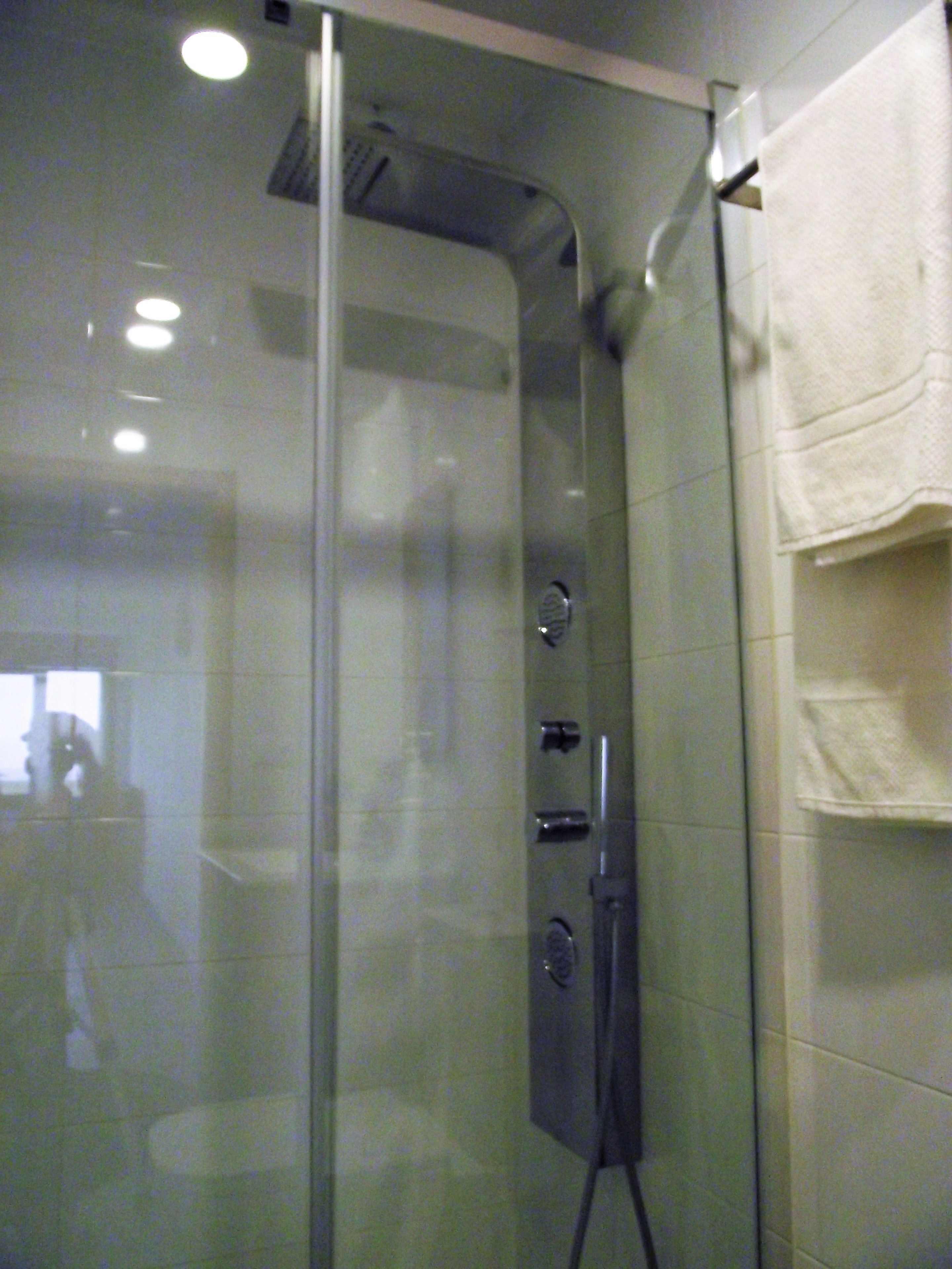 Ducha con cerramiento de vidrio, panel de control, perchero y el reflejo visible de una persona.
