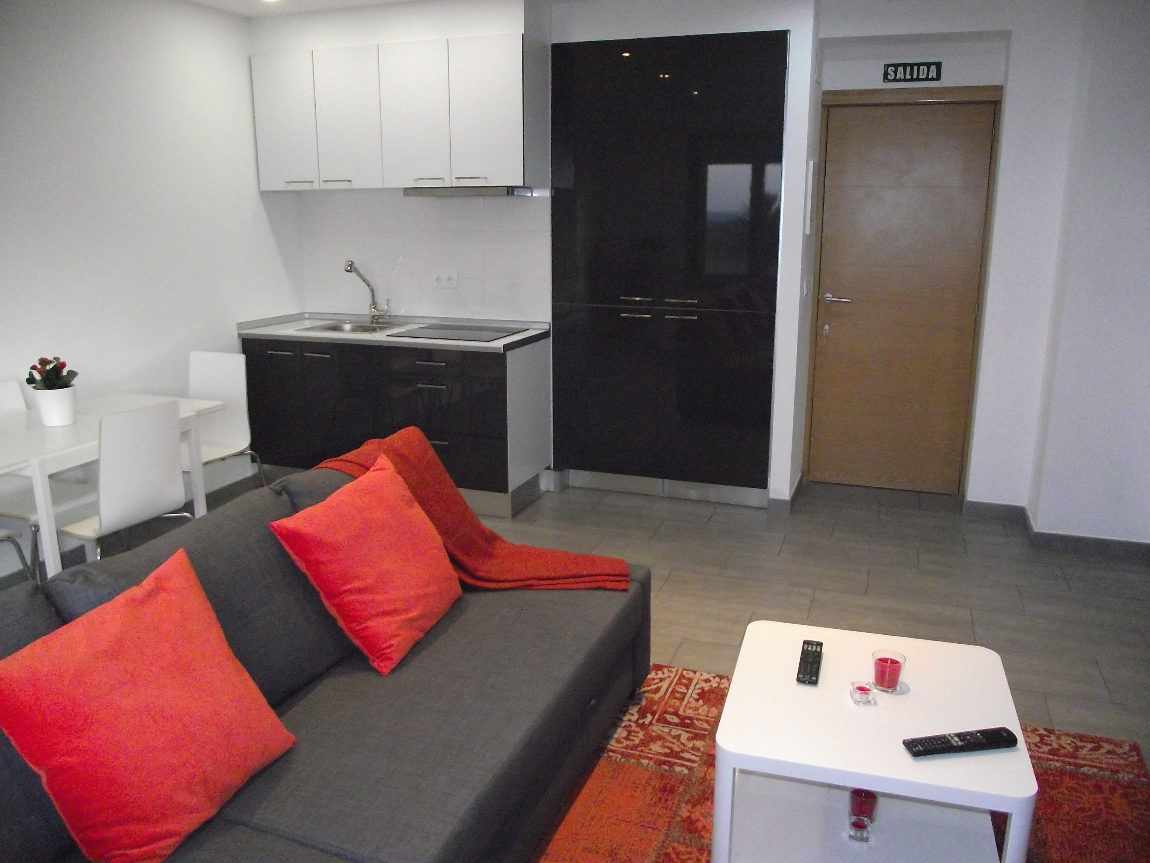 Una sala moderna con un sofá gris, cojines rojos, una mesita blanca con un control remoto, y una zona de cocina con armarios blancos y una puerta negra.