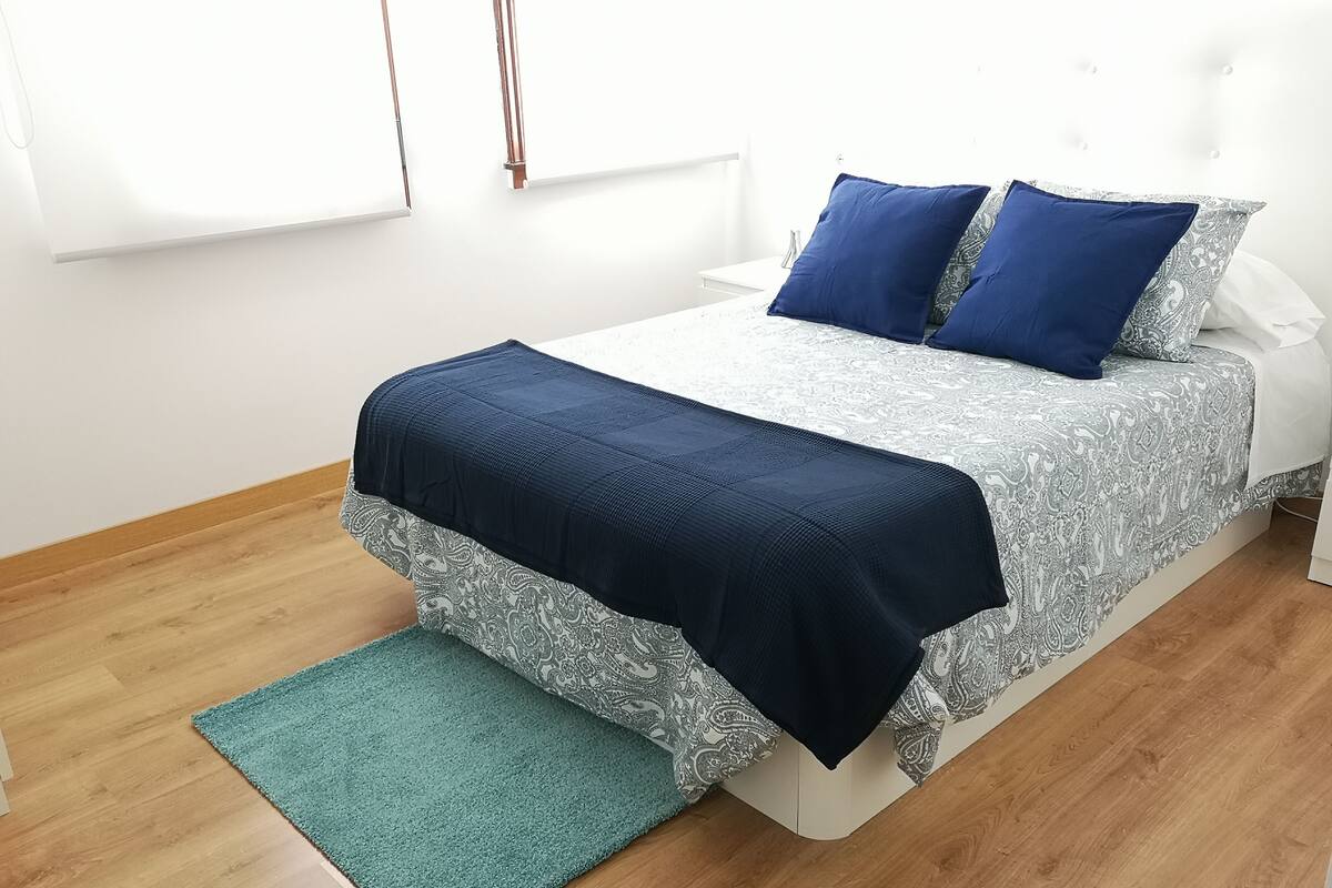 Una cama bien hecha con una colcha estampada y dos almohadas, una alfombra verde pequeña y una mesita de noche blanca.