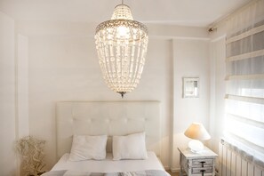 A bedroom with a large, ornate chandelier, a tufted headboard, a bedside table with a lamp, and a view of the outside through sheer curtains.