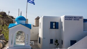A building with a sign reading "MYKONOS PRO-CARE HOLIDAY SUITES" and a flag.