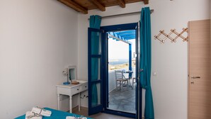A room with a bed, a desk with a lamp, a chair, and a view of the sea through an open door.