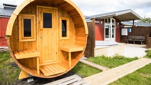 Un bain à remous en bois, doté d’une porte et de fenêtres, entouré d’une terrasse en bois et d’un bâtiment à l’extérieur rouge.
