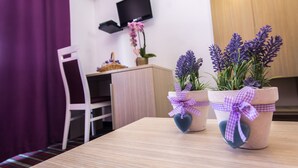 A room with a wooden table, two potted plants with purple flowers, a white chair, a television mounted on the wall, and a purple curtain. - Hotel Villa Sandi (Cavle)