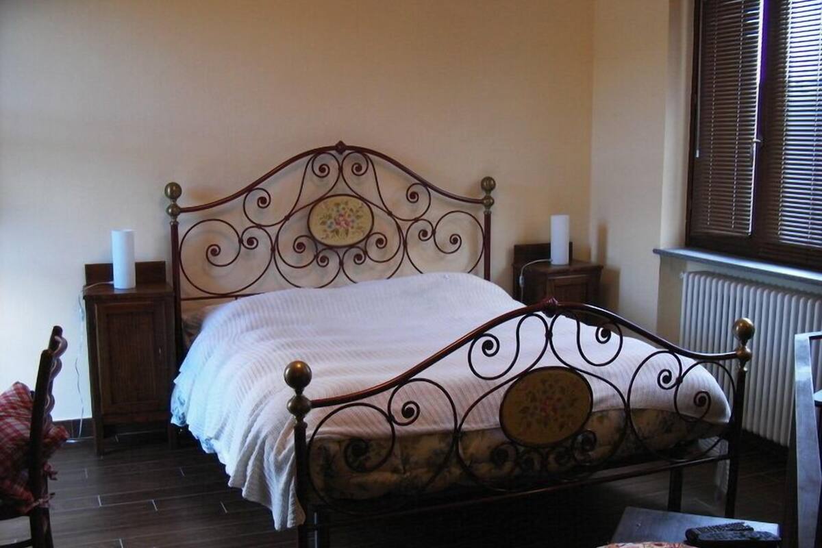 A bedroom with a metal-framed bed, a wooden nightstand, a chair, and a window with blinds.