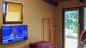 A room with a television displaying a news program, a red metal storage unit, a window with blinds, and a door leading outside. - Residence delle Rose (Monforte d'Alba)