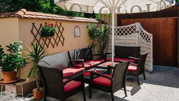 An outdoor patio with wicker chairs and red cushions, a large umbrella, and potted plants.