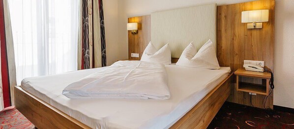A hotel room with a large bed, two bedside lamps, a wooden headboard, and a small bedside table with a phone.