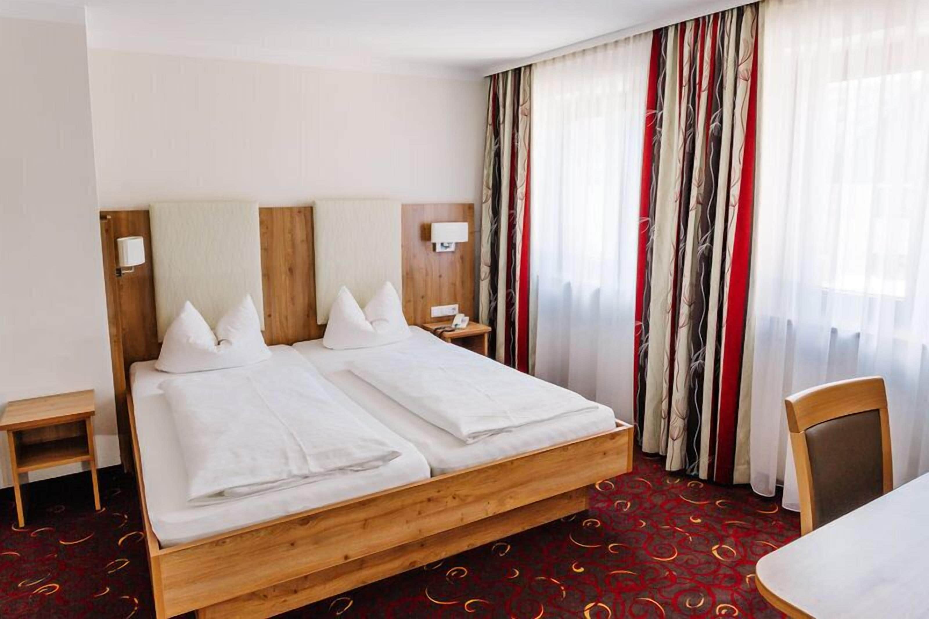 Una cama doble con sábanas blancas, cabecera de madera, mesita de noche con lámpara y ventana con cortinas translúcidas.
