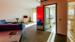 A modern hotel room with a sofa, a small table with a book, a red accent wall, and a kitchen area visible through a glass door. - Welc-oM Manuela (Selvazzano Dentro)