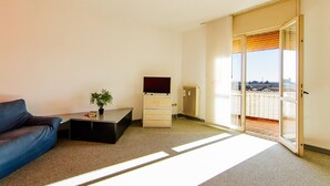 A modern hotel room with a blue sofa, a small table with a plant, a TV, and a sliding glass door leading to a balcony with a city view. - Welc-oM Manuela (Selvazzano Dentro)