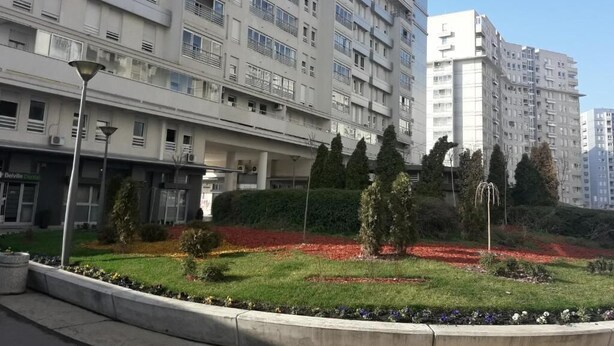 En flervåningsbyggnad med bostäder, balkonger och en vacker trädgård med röda blommor, under en klar himmel.