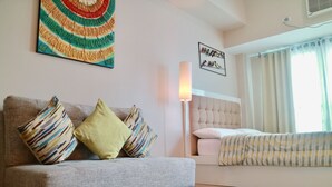 A modern living room with a grey sofa, colorful cushions, a tall floor lamp, a bed with striped bedding, and a large window with curtains. - Eastwood Le Grand Tower 3 (Quezon City)