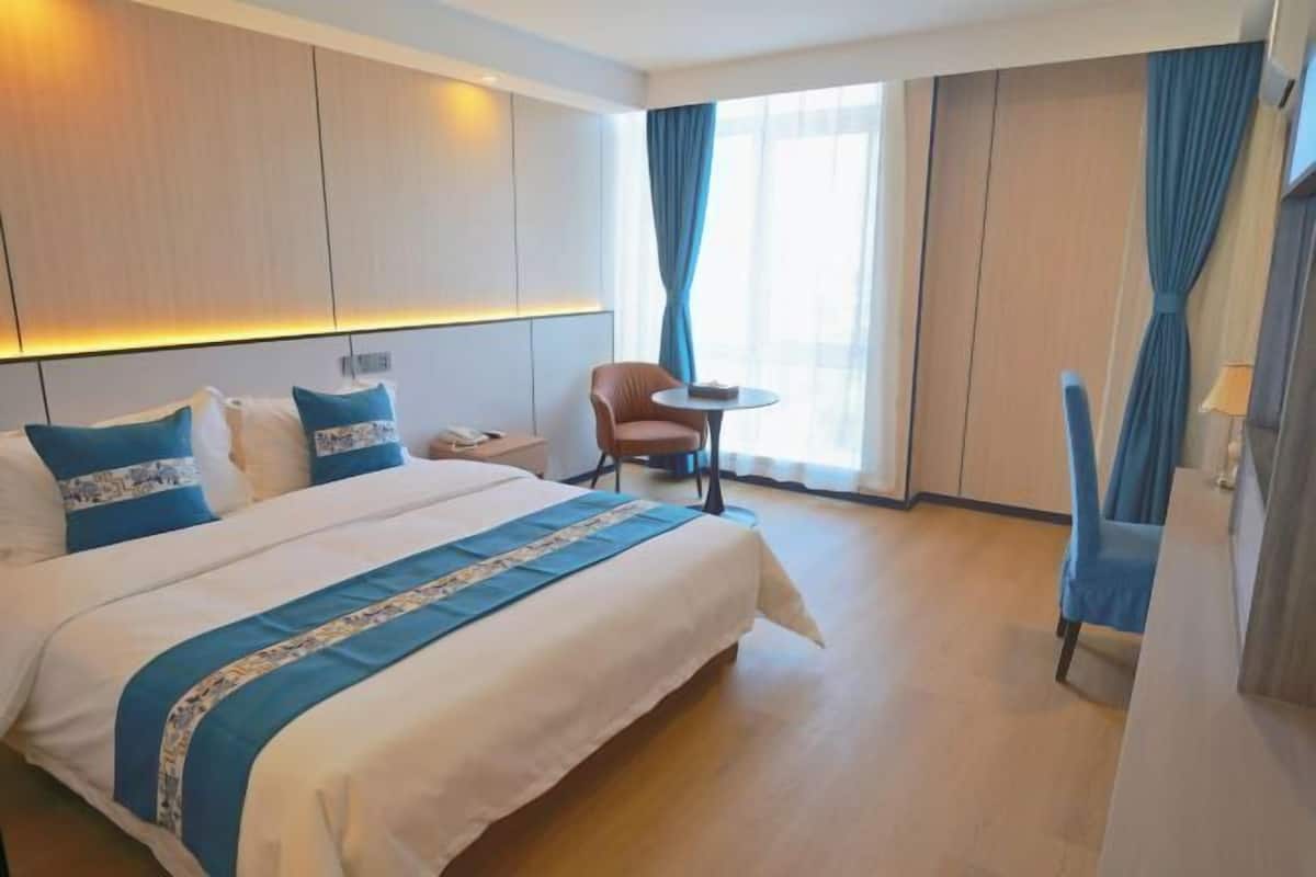 A hotel room with a large bed, a small table, a chair, and a window with curtains.
