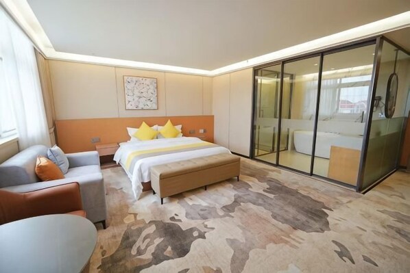 A hotel room with a bed, a sofa, a desk, a glass-enclosed bathroom, and a large window.