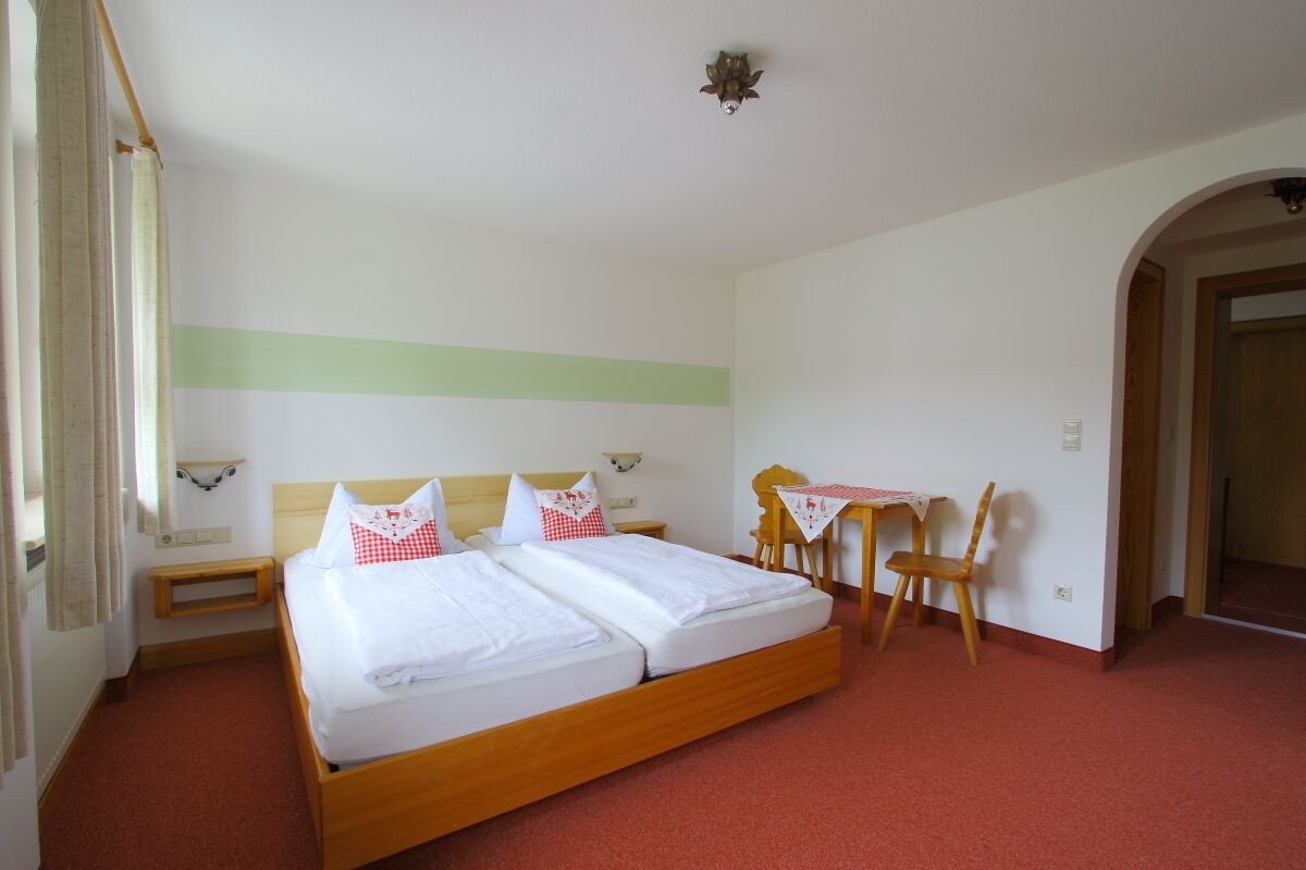 A double bed with white bedding and red and white pillows, a wooden nightstand, a small table with a tablecloth, and a chair.