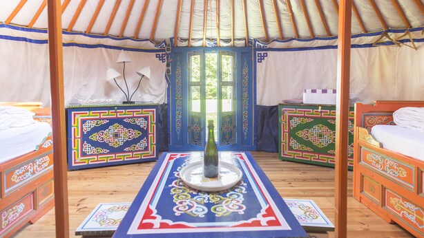 Une yourte traditionnelle avec deux lits, une table au centre sur laquelle se trouve une bouteille, et des motifs décoratifs sur les murs et le sol.